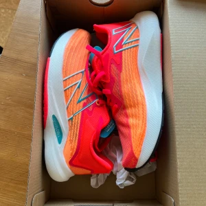 Orange och röda sneakers från New Balance - Snygga och färgglada sneakers från New Balance i en mix av orange och rött med vita och svarta detaljer. Skorna har snörning och en bekväm sula, perfekt för träning eller vardagsbruk.