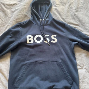 Mörkblå hoodie från Boss - Säljer en snygg mörkblå hoodie från Boss med stor vit logga på bröstet. Tröjan har en klassisk känguruficka och justerbar huva med snören. Perfekt för en avslappnad stil.