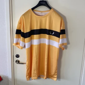 Gul och svart randig t-shirt från Fred Perry - Helt ny . Snygg gul t-shirt från Fred Perry med svarta och vita ränder över bröstet. Den har en normal passform och korta ärmar, perfekt för en avslappnad stil. Den ikoniska Fred Perry-loggan är broderad på bröstet.