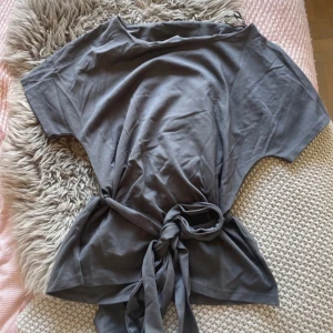 Grå kortärmad topp med knytband - Säljer en stilren grå topp med korta ärmar och knytband i midjan. Perfekt för en avslappnad look. Passar bra till både jeans och kjol.
