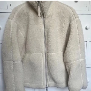 Beige teddyjacka med dragkedja - Mysig beige teddyjacka med dragkedja framtill. Jackan har en hög krage och långa ärmar, perfekt för kyliga dagar. Den fluffiga texturen ger en trendig look och den neutrala färgen gör den lätt att matcha med olika outfits.