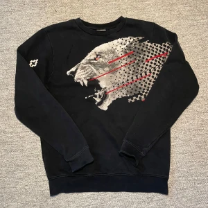 Svart tröja med tryck från Marcelo Burlon - Säljer en svart tröja från Marcelo Burlon med ett coolt tryck av ett djurhuvud i grått och rött. Tröjan har långa ärmar och är tillverkad i bomull för en bekväm känsla. Perfekt för dig som vill ha en unik stil!