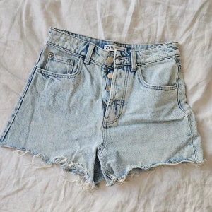 Ljusa jeansshorts från Zara - Säljer ett par ljusa jeansshorts från Zara med hög midja och fransiga kanter. Shortsen har en knappgylf med tre knappar och klassiska fem fickor. Perfekta för sommardagar!