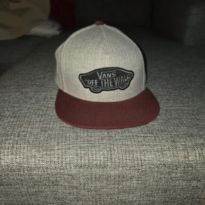 Grå och vinröd keps från Vans - Snygg keps från Vans i grått och vinrött med klassisk 'Off The Wall'-logga framtill. Kepsen har en justerbar rem baktill för perfekt passform. Perfekt för en avslappnad stil.