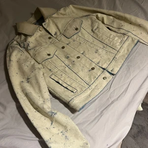 Beige jeansjacka från Eytys - Säljer en unik beige jeansjacka från Eytys med blå stänkdetaljer. Jackan har en klassisk krage, knappar framtill och två bröstfickor med lock. Insidan är fodrad med blått tyg som ger en snygg kontrast. Perfekt för dig som vill sticka ut!