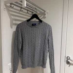 Grå stickad tröja från Gant - Säljer en stilren grå stickad tröja från Gant med kabelstickat mönster. Tröjan har långa ärmar och en rund halsringning. Perfekt för kyligare dagar. 🧶