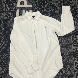Ralph Lauren skjorta - Klassisk vit skjorta från Ralph Lauren med broderad logotyp på bröstet. Skjortan har långa ärmar och knappar framtill. Perfekt för en stilren look. 