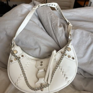 Vit axelväska med kedjedetaljer - Snygg vit axelväska med unika kedjedetaljer och dekorativa sömmar. Väskan har en dragkedja upptill och en justerbar axelrem med nitar. Perfekt för att ge din outfit en edgy touch.