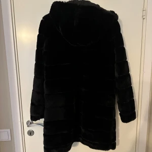 Svart pälsjacka med huva - Snygg och elegant svart pälsjacka med huva och dragkedja. Perfekt för kalla dagar när du vill hålla dig varm och stilren. Jackan har långa ärmar och en mjuk, lyxig känsla.