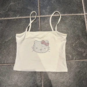 Hello kitty linne  - Säljer en söt vit linne med smala axelband och ett glittrigt strassmotiv framtill. Perfekt för en avslappnad look med en touch av bling. Passar bra till jeans eller shorts!