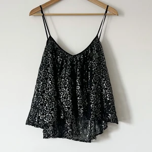Svart glittrig linne - Snygg svart linne med glittrigt mönster och tunna axelband. Perfekt för en kväll ute eller festliga tillfällen. Den har en lös passform och en v-ringad design som ger en elegant look.