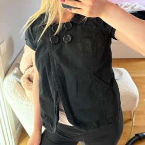 Svart kortärmad jacka med knappar - Snygg svart kortärmad jacka med stora knappar framtill. Perfekt för en stilren look. Jackan har en klassisk krage och två fickor fram.💕💕💕