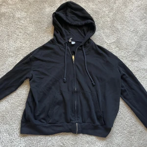 Svart hoodie från H&M - Säljer en klassisk svart hoodie från H&M Divided. Den har en dragkedja framtill och en praktisk huva med dragsko. Perfekt för en avslappnad stil 💕