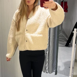 Beige teddyjacka - Mysig beige teddyjacka med knappar framtill. Perfekt för kyliga dagar med sin fluffiga och varma design. Jackan har en avslappnad passform och långa ärmar.