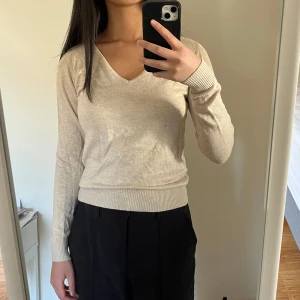 Beige v-ringad tröja - Säljer en stilren tunn beige tröja med v-ringning och långa ärmar. Tröjan har ribbade muddar vid ärmslut och nederkant, vilket ger en snygg passform. Perfekt för en avslappnad look.
