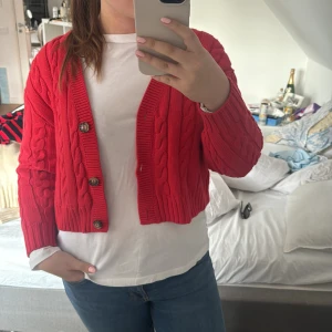 Röd stickad kofta från Monki - Säljer en snygg röd stickad kofta från Monki med flätstickat mönster och stora knappar. Koftan är croppad och har långa ärmar, perfekt för att ge en färgklick till din outfit. Passar bra till både jeans och kjol. 🌟för liten på mig och aldeig använd! Perfekt till sommaren ❤️