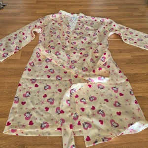 Beige pyjamasöverdel & shorts från Snoopy - Säljer en söt pyjamas/morgonrock med Snoopy-motiv och rosa hjärtan, samt ett par matchande shorts. Pyjamasen har långa ärmar och knytband i midjan. Shortsen har både resår och knappar i sidan. Perfekt för en mysig hemmakväll! 💖 Oanvända. Se mina andra annonser och köp bundle med 10% rabatt och samfrakt.