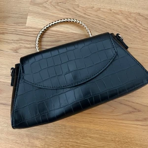 Svart handväska med krokodilmönster - Snygg svart handväska med krokodilmönster och ett elegant flätat handtag i metall. Perfekt för att ge din outfit en stilfull touch. Väskan har en unik form och stängs med en lock.