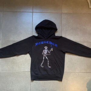 Svart hoodie från Detached med skelettmotiv - Storlek S och jag är 173 cm lång. Säljer en svart hoodie från Detached med ett coolt skelettmotiv och blå text på bröstet. Tröjan har en klassisk känguruficka och en bekväm huva. Perfekt för en avslappnad stil.