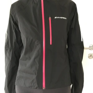 Svart cykel regn jacka Everest stl 36 Ny - Svart regn jacka cykel jacka från Everest med rosa detaljer. Jackan har en dragkedja framtill och en bröstficka med dragkedja. Det finns en reflexrand på ryggen och ärmarna. En bröstficka med dragkedja och en liten ficka med dragkedja nedtill på ryggen. Extra lång i bak vid cykling. Extra långa ärmar vid cykling. Stl 36. Nylon. Ny aldrig använd. En defekt, hängaren som varit limmad i nackens insida har lossnat, inget som påverkar funktion eller utseende. Vattenpelare 10 000.  Nypris: 899kr