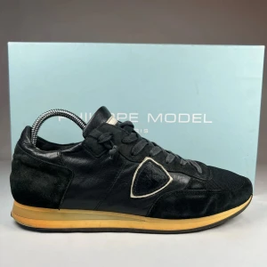 Svarta sneakers från Philippe Model - Snygga svarta sneakers från Philippe Model med en stilren design. Skorna har snörning och en kontrasterande gul sula som ger en cool touch. Tillverkade i en kombination av skinn och mocka för en lyxig känsla. Perfekta för en trendig look.