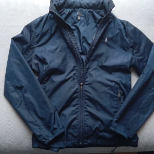 jacka från Polo Ralph Lauren - Säljer en stilren mörkblå vindjacka från Polo Ralph Lauren. Jackan har dragkedja framtill och en broderad logotyp på bröstet. Perfekt för blåsiga dagar och ger en klassisk look.den är en tjockare vind jacka som tål kyla. 