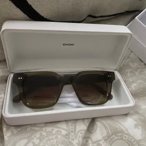 Solglasögon från Chimi - Snygga solglasögon från Chimi i en stilren fyrkantig design. Glasögonen har en mörkgrön färg och kommer i ett elegant vitt fodral. Perfekta för att ge din outfit en cool touch. Pris kan diskuteras. Använda en gång bara, då jag inte tycker de är riktigt min stil längre.