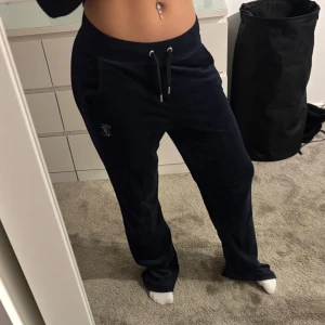 Mörkblå mjukisbyxor från Juicy Couture - Säljer ett par mörkblå mjukisbyxor från Juicy Couture med snörning i midjan och glittrig text på baksidan. Byxorna har en avslappnad passform och är perfekta för en bekväm stil.