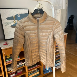 Beige quiltad jacka från Vero Moda - Snygg beige quiltad jacka från Vero Moda med dragkedja framtill. Jackan har en stilren design och är perfekt för kyligare dagar. Den är tillverkad i polyester och har långa ärmar.