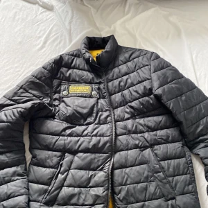 Svart quiltad jacka från Barbour - Säljer en svart quiltad jacka från Barbour International med gul insida. Jackan har en dragkedja framtill och en bröstficka med Barbour-logga. Perfekt för kyligare dagar!