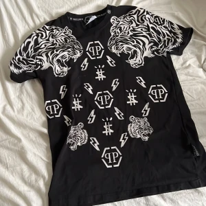 ÄKTA Svart t-shirt med tigertryck från Philipp Plein - Cool svart t-shirt från Philipp Plein med ett iögonfallande vitt tigertryck på ärmarna och flera logotyper och blixtar på framsidan. Perfekt för dig som vill sticka ut med en unik stil.