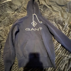 Mörkblå hoodie från GANT - Säljer en snygg mörkblå hoodie från GANT med logga framtill. Den har en klassisk känguruficka och justerbar huva med dragsko. Perfekt för en avslappnad stil.