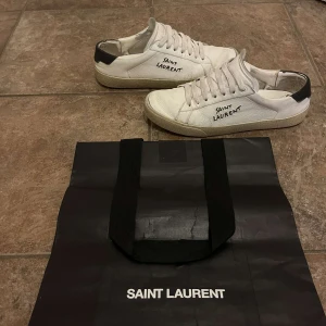 Saint laurent vita sneakers - Snygga vita sneakers från Saint Laurent med svart detalj på hälen och handskriven logga på sidan. Skorna har en klassisk design med snörning och en bekväm sula. Perfekta för en stilren look.