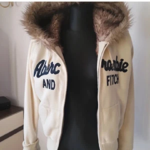Beige hoodie från Abercrombie & Fitch - Säljer en beige hoodie från Abercrombie & Fitch med pälskantad huva. Den har dragkedja framtill som kan krångla ibland och ribbade muddar vid ärmslut och nederkant. 💕Pris går att diskutera 