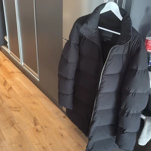 Svart pufferjacka från Emporio Armani - Säljer en stilren svart pufferjacka från Emporio Armani. Jackan är tillverkad i komfortabel stretchpolyester och har en dragkedja framtill. Perfekt för kyligare dagar med sin långa design och vadderade stil. Ordinare pris 5999