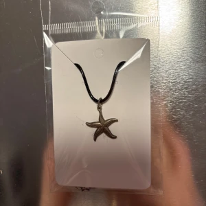 Halsband med sjöstjärna - Snyggt halsband med en silverfärgad sjöstjärna som hänge. Hängsmycket sitter på ett svart band som ger en stilren look. Perfekt för att lägga till en marin touch till din outfit.
