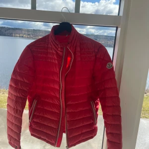 Röd dunjacka från Moncler - Snygg röd dunjacka från Moncler med dragkedja och hög krage. Jackan har långa ärmar och två fickor med dragkedja framtill. Perfekt för kyligare dagar. Köpt från VonGreven Closet för 3600kr