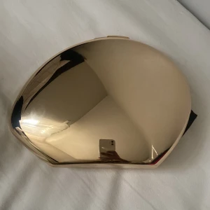 Guldig kuvertväska - Elegant guldig kuvertväska med en stilren design. Väskan har en metallisk yta och en kedja som axelrem. Perfekt för att ge en lyxig touch till din outfit.