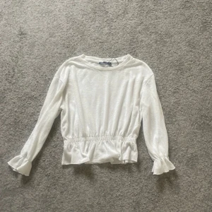 Vit tröja från Zara - Säljer en stilren vit tröja från Zara med långa ärmar och elastisk midja. Tröjan har volangdetaljer vid ärmslut och nederkant, vilket ger en söt touch.