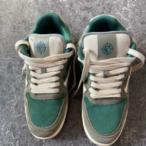 Gröna och beige sneakers med logotyp - Snygga sneakers i grönt och beige med en unik logotyp på plösen och hälen. Skorna har en klassisk snörning och en bekväm sula i gummi. Perfekta för en avslappnad stil.