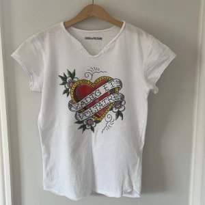 Vit t-shirt med hjärtmotiv från Zadig & Voltaire - Säljer en cool vit t-shirt från Zadig & Voltaire med ett stort hjärtmotiv och blommor på framsidan. T-shirten har korta ärmar och en rund halsringning med råa kanter. Frakt ingår inte i priset!