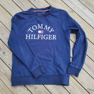 Blå tröja från Tommy Hilfiger - Säljer en snygg blå tröja från Tommy Hilfiger med deras ikoniska logga tryckt på framsidan. Tröjan har långa ärmar och ribbade muddar vid ärmslut och nederkant. Perfekt för en stilren och avslappnad look.
