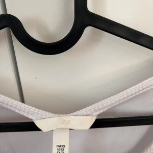 Vit ribbad topp från H&M - Säljer en vit ribbad topp från H&M med en snygg knytdetalj framtill. Toppen har långa ärmar och är tillverkad av återvunnen polyester och LivaEco viskos. Perfekt för en stilren look. Bodyn är helt ny och aldrig använd. 