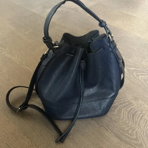 Mörkblå zara skinnväska med axelrem - Snygg mörkblå skinnväska med justerbar axelrem och praktisk dragskoöppning. Perfekt för att bära dina dagliga nödvändigheter med stil. Väskan har en rymlig interiör och en elegant design.