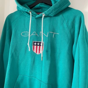 Turkos/grön hoodie från GANT - Säljer en snygg turkos hoodie från GANT med broderad logga och emblem på bröstet. Hoodien har en klassisk känguruficka och justerbar huva med vita snören. Perfekt för en avslappnad stil. Nypris 1600kr. Obs. Ena metallgrejen på snöret har släppt därav priset.