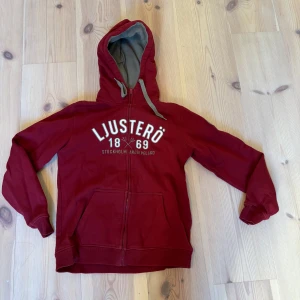 Röd hoodie med tryck - Säljer en röd hoodie med tryck 'Ljusterö 1869 Stockholm Norrköping' på framsidan. Den har en dragkedja och en justerbar huva med grå insida. Perfekt för en avslappnad stil.