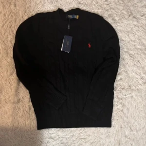 Svart stickad tröja från Polo Ralph Lauren - Säljer en  väldigt ren  svart stickad tröja från Polo Ralph Lauren med det klassiska röda logotypbroderiet på bröstet. Tröjan har en rund halsringning och långa ärmar med ribbade muddar. Perfekt för en elegant och avslappnad look. 