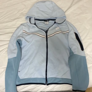 Ljusblå hoodie från Nike - Säljer en ljusblå hoodie från Nike med dragkedja och huva. Jackan har en mörkare blå kant och en ficka med dragkedja på ärmen. Perfekt för en sportig look eller en avslappnad dag.