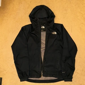 Svart vindjacka från The North Face - Säljer en svart vindjacka från The North Face med huva och dragkedja. Jackan har en stilren design och är perfekt för blåsiga dagar. Den är lätt och smidig att bära med sig. Passar bra för utomhusaktiviteter och vardagsbruk.