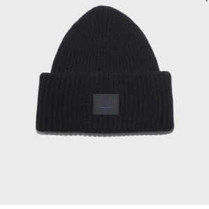 Svart ribbad mössa från Acne Studios - Snygg svart ribbad mössa från Acne Studios med ett diskret broderat ansikte på framsidan. Perfekt för att hålla värmen under kyliga dagar. En stilren accessoar som passar till det mesta.